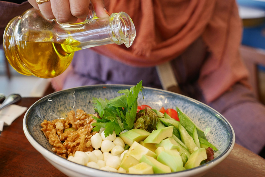 A person pouring golden olive oil from a glass carafe onto a fresh salad bowl containing walnuts, avocado, mozzarella pearls, and greens.