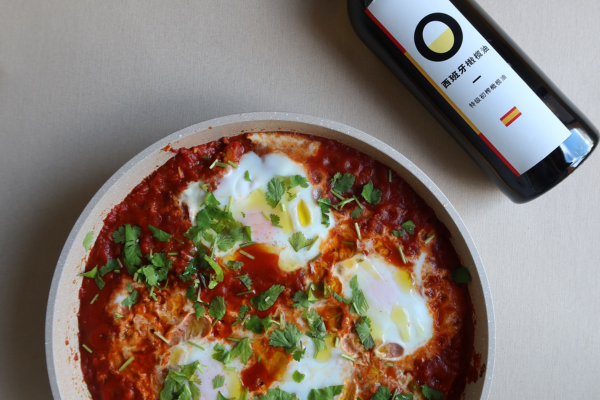 A top-down view of a shakshuka dish with poached eggs and fresh herb in a beige pan, placed next to a bottle of Spanish olive oil.